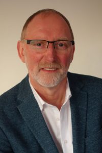 Porträt eines älteren Mannes mit Brille, der in einem blauen Blazer gekleidet ist. Er lächelt freundlich und hat einen gepflegten Bart. Der Hintergrund ist neutral gehalten.
