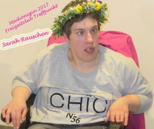 Junger Mann mit einem Blumenkranz auf dem Kopf, der entspannt in einem Stuhl sitzt und ein graues T-Shirt mit der Aufschrift "CHIC" trägt. Hintergrund ist schlicht und hell.
