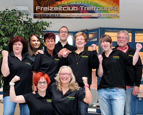 Teamfoto einer Gruppe von Mitarbeitern in schwarzen Poloshirts, die vor einem bunten Wandbild mit Regenbogen stehen. Die Gruppe zeigt eine positive Stimmung mit verschiedenen Gesten und Lächeln. Ideal für die Präsentation von Teamarbeit und Gemeinschaftsgefühl.