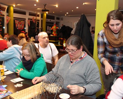 Leute sitzen an Tischen. Einige spielen ein Spiel mit vielen kleinen Teilen. Es gibt Essen und Getränke auf den Tischen.