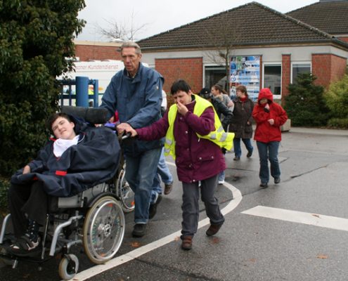 Eine Gruppe von Kindern und Erwachsenen, darunter ein Junge im Rollstuhl, geht auf einem Gehweg. Ein Mann schiebt den Rollstuhl, während andere in bunten Jacken hinterherlaufen. Im Hintergrund sind Gebäude und ein bewölkter Himmel zu sehen.