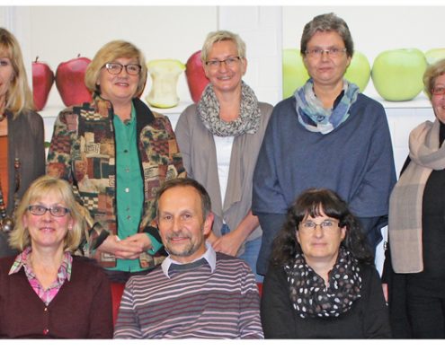 Gruppe von acht Personen, die in einem freundlichen Umfeld posieren, mit einer bunten Dekoration im Hintergrund.
