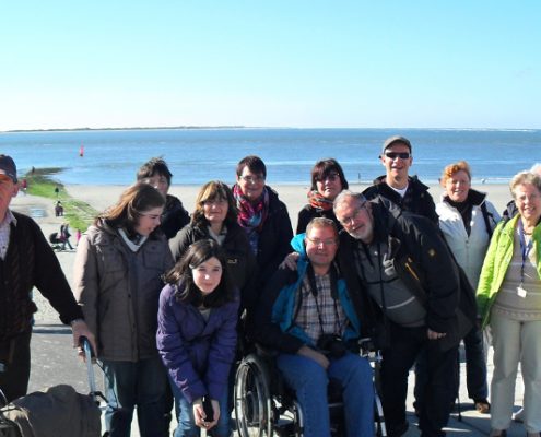 Eine Gruppe von Menschen steht am Strand. Einige haben Rollatoren oder Gehhilfen. Im Hintergrund ist das Meer zu sehen.