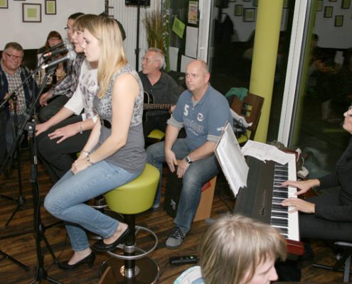 Bandaufführung in einem gemütlichen Lokal, mit einer Sängerin, die am Mikrofon steht, während Musiker im Hintergrund Klavier und andere Instrumente spielen. Die Atmosphäre ist entspannt und lebhaft.
