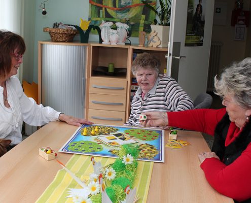 Drei ältere Menschen spielen ein Brettspiel. Ein Hund sitzt daneben. Es ist ein gemütliches Zimmer mit Blumen auf dem Tisch.
