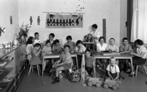 Kinder sitzen an Tischen in einem Raum. Es gibt Spielsachen auf dem Tisch und auf dem Boden.