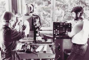 Historisches Schwarz-Weiß-Foto von zwei Personen in einem Raum mit technischen Geräten, darunter ein Plattenspieler und ein Mischpult. Die Szene vermittelt eine Atmosphäre der frühen Audioaufnahme-Technologie.