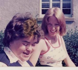 Zwei Personen stehen draußen. Eine Frau und ein Mann lächeln. Sie stehen vor einem grünen Busch.