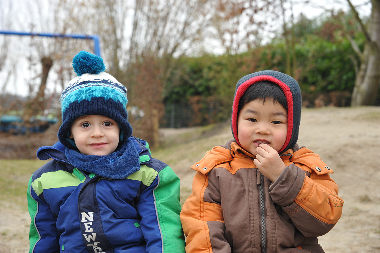 Zwei fröhliche Kinder im Winter, eines trägt eine blaue Mütze und einen blauen Mantel, das andere eine rote Mütze und einen orangenen Mantel, sitzen im Freien vor einem winterlichen Hintergrund.