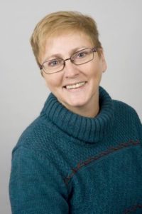 Eine Frau lächelt in die Kamera. Sie trägt eine blaue Jacke und eine Brille.