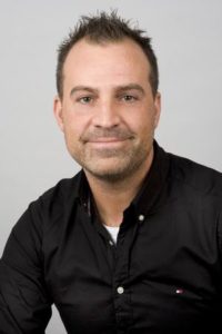 Porträt eines Mannes mit kurzem Haar, lächelnd, in einem schwarzen Hemd vor einem neutralen Hintergrund.