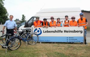 Eine Gruppe von Kindern mit Fahrrädern und Helmen steht vor einem Van. Ein Mann steht daneben.