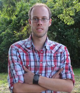 Junger Mann mit Brille und kariertem Hemd steht mit verschränkten Armen vor einer grünen Gartenlandschaft.