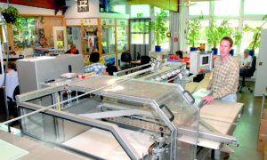 Werkstatt mit Maschinen und Personen. Ein Mann steht neben einer Maschine. Es gibt viele Geräte und Werkzeuge.