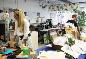 Frau arbeitet an einem Tisch in einem kreativen Raum, umgeben von Bastelmaterialien und Pflanzen, während im Hintergrund weitere kreative Aktivitäten stattfinden.