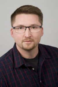Ein Mann mit Brille lächelt. Er trägt ein kariertes Hemd und ein schwarzes T-Shirt.