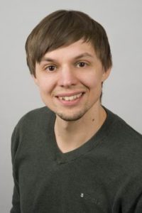 Ein Mann lächelt in die Kamera. Er hat kurze, braune Haare und trägt ein grünes Shirt.