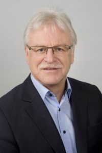 Ein älterer Mann mit Brille und Bart. Er trägt ein blaues Hemd und eine Jacke.