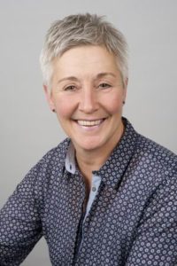 Porträt einer lächelnden älteren Frau mit kurzen grauen Haaren, die ein kariertes Hemd trägt. Der Hintergrund ist neutral und hell, was die freundliche Ausstrahlung unterstreicht.