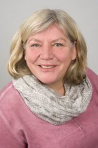 Porträt einer lächelnden Frau mit blonden Haaren, die einen grauen Schal trägt. Sie hat einen freundlichen Ausdruck und steht vor einem neutralen Hintergrund.