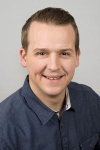 Professionelles Porträt eines lächelnden Mannes in einem blauen Hemd vor neutralem Hintergrund. Ideal für Unternehmensprofile oder persönliche Webseiten.