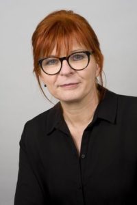 Professionelle Frau mit roten Haaren und Brille, die in die Kamera lächelt, vor neutralem Hintergrund.