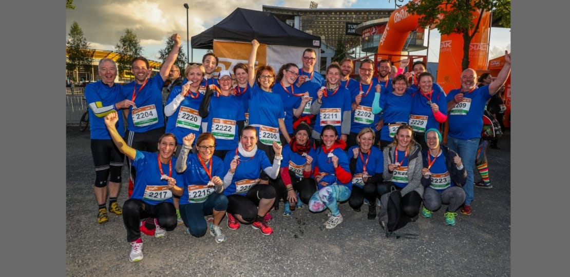Gruppenselfie von Teilnehmern eines Sportevents, die in blauen T-Shirts gekleidet sind und stolz ihre Medaillen zeigen. Die Atmosphäre ist festlich, mit einem Zielbogen im Hintergrund.