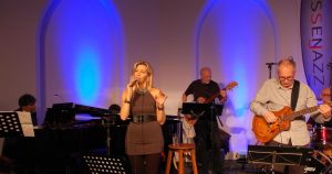 Eine Frau singt auf einer Bühne. Sie steht vor einer Band mit Gitarre und Klavier. Es ist dunkel und hat viele Lichter.