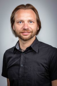 Ein Mann lächelt in die Kamera. Er trägt ein schwarzes Hemd mit einer weißen Tasche auf der Brust.