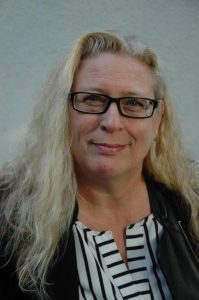 Porträt einer Frau mit langen blonden Haaren und Brille, die freundlich in die Kamera lächelt. Sie trägt eine schwarze Jacke und hat einen neutralen Hintergrund.