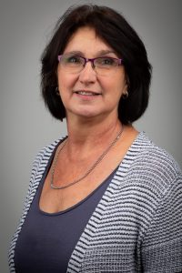 Angelika Schrimpf Porträt einer Frau mittleren Alters mit dunklen Haaren und einer Brille, die ein gestreiftes Oberteil trägt. Der Hintergrund ist neutral und unauffällig.