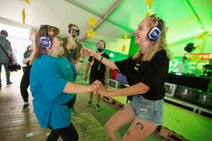 Zwei Frauen tanzen fröhlich auf einer Silent Disco, während sie Kopfhörer tragen. Im Hintergrund sind weitere Personen zu sehen, die ebenfalls an der Veranstaltung teilnehmen. Die Atmosphäre ist lebhaft und bunt, mit grünen Zeltwänden.