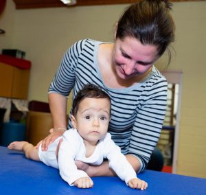 Eine Mutter unterstützt ihr Baby beim Spielen auf einer blauen Matte in einem Raum für frühkindliche Entwicklung. Das Baby liegt auf dem Bauch und schaut neugierig in die Kamera.