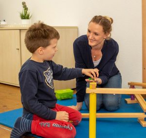 Eine Lehrerin spielt mit einem Jungen in einem Lernraum. Sie interagieren spielerisch mit einem Holzspielzeug auf einer blauen Matte. Im Hintergrund sind Regale und Pflanzen sichtbar.
