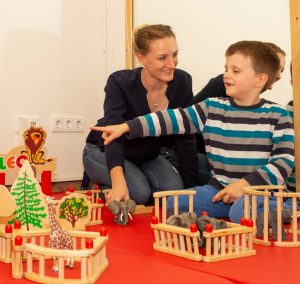 Eine Mutter und ihr Sohn spielen gemeinsam mit Holzspielzeug auf einem roten Teppich. Der Junge zeigt auf eine Figur, während sie ein Spielzeughaus und eine Weihnachtsbaum-Dekoration aufgebaut haben.
