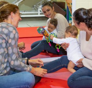 Zwei Mütter spielen mit ihren Kleinkindern auf einer roten Matte, während sie Spielzeug verwenden. Die Atmosphäre ist freundlich und fördert die Entwicklung der Kinder durch interaktives Spiel.