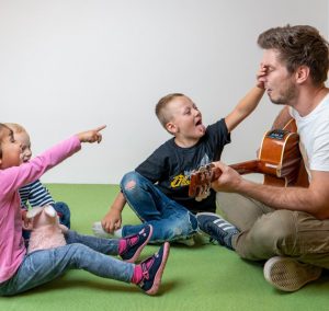 Ein Musikpädagoge spielt Gitarre, während zwei Kinder begeistert mit ihm interagieren. Ein Mädchen in rosa zeigt auf ihn, während ein Junge lacht und mitmacht. Die Szene findet in einem hellen Raum mit grünem Boden statt.