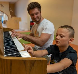 Ein Lehrer zeigt einem Schüler, wie man Klavier spielt, während sie gemeinsam an einem Klavier sitzen. Der Schüler konzentriert sich auf die Tasten, während der Lehrer ihn anleitet.