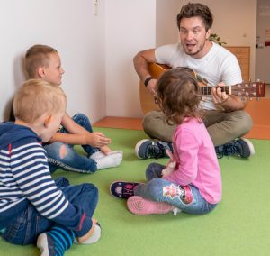 Ein Mann spielt Gitarre. Zwei Kinder sitzen auf dem Teppich und hören zu.