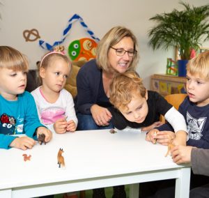 Drei Kinder spielen mit Tieren auf einem Tisch. Eine Frau sitzt daneben und schaut zu.