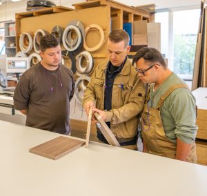 Drei Handwerker betrachten gemeinsam ein Holzprojekt in einer Werkstatt, umgeben von Werkzeugen und Materialien.