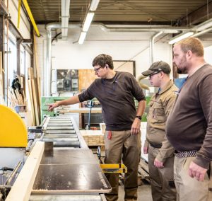 Drei Handwerker arbeiten an einer Holzverarbeitungsmaschine in einer Werkstatt, während sie die Bedienung und Techniken zur Holzverarbeitung erlernen.