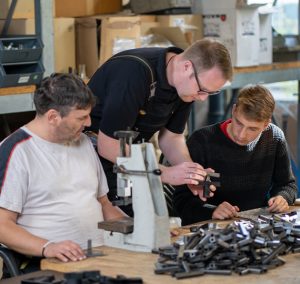 Zwei Männer arbeiten an Metallteilen. Einer hält ein Werkzeug, der andere schaut hin.
