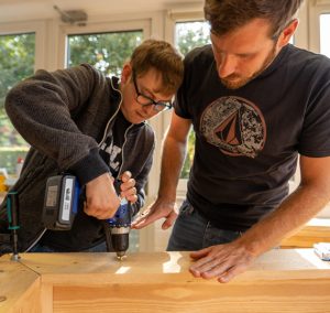 Zwei Männer arbeiten gemeinsam an einem Holzprojekt, einer benutzt einen Akkuschrauber, während der andere das Werkstück hält. Helle Umgebung mit großen Fenstern, die natürliches Licht hereinlassen.