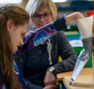 Eine Lehrerin unterstützt eine Schülerin beim Lernen in einem Klassenzimmer, während andere Schüler im Hintergrund beschäftigt sind.