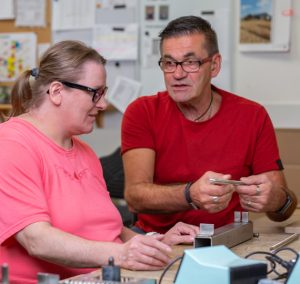 Zwei Personen, eine Frau in einem rosa T-Shirt und ein Mann in einem roten T-Shirt, sitzen an einem Tisch und diskutieren. Auf dem Tisch liegen verschiedene Materialien und Unterlagen. Die Szene vermittelt eine freundliche und kooperative Atmosphäre.