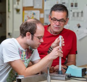 Zwei Männer führen Experimente in einem Labor durch, wobei einer einen Messzylinder hält und der andere aufmerksam zusieht. Im Hintergrund sind Laborgeräte und Notizen sichtbar.