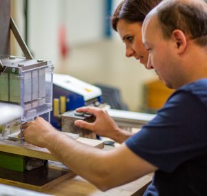 Zwei Personen arbeiten konzentriert an einem technischen Projekt in einer Werkstatt, während sie Materialien und Werkzeuge verwenden.