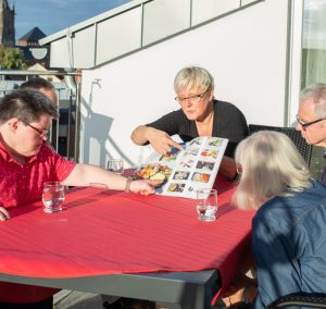 Menschen sitzen an einem Tisch. Eine Frau hält ein Buch. Es sind mehrere Personen zu sehen.