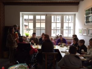 Gruppe von Menschen, die an einem Tisch in einem hellen Raum mit Fenstern zusammen sitzen und diskutieren, umgeben von Pflanzen und einer einladenden Atmosphäre.